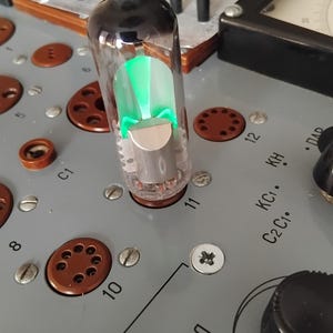 Peut inclure: Gros plan d'un appareil électronique vintage avec une lampe à vide allumée. Le tube transparent émet une lumière verte, sur un panneau gris avec des cadrans, des boutons et d'autres composants. Le mot "НАКАЛ" est visible.
