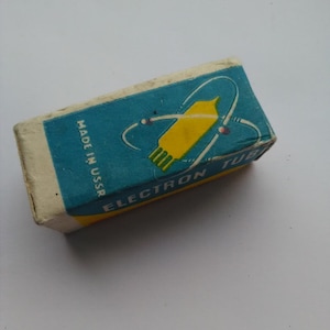 Può includere: Una scatola rettangolare vintage con la parte superiore blu e una grafica gialla e verde di un tubo elettronico. La scatola ha la scritta "ELECTRON TUBE" e "MADE IN USSR". La scatola è fatta di cartone e ha un aspetto usurato.