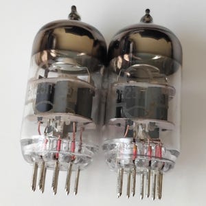 May include: Two vintage vacuum tubes with clear glass bulbs and dark grey tops. Each tube has internal components and a series of pins at the base. These tubes are likely used in electronic devices.