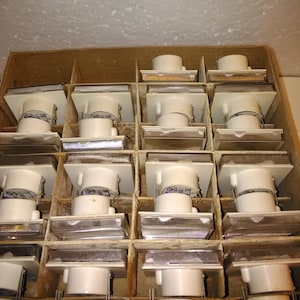 May include: A wooden box containing numerous white and clear light fittings. Each fitting features a square top, a cylindrical body, and a rectangular base with a clear, textured lens. The fittings are organised in separate compartments.