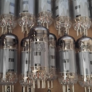 Peut inclure: Une collection de tubes à vide vintage, chacun avec un boîtier en verre et une base en métal. Les tubes sont disposés en rangées, avec des composants internes visibles. Certains tubes portent des inscriptions, dont "6K4" et "W075".