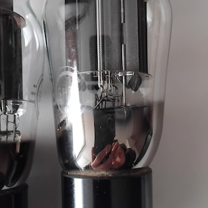 Puede incluir: Dos válvulas de vacío vintage con bombillas de vidrio transparente y bases negras. Los componentes internos son visibles, incluyendo estructuras metálicas y filamentos. Los tubos están diseñados para dispositivos electrónicos y tienen múltiples pines en la base.