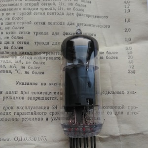 Può includere: Una valvola termoionica vintage con una parte superiore nera e un corpo in vetro trasparente, che rivela i componenti interni. Il tubo ha più pin alla base ed è posto su uno sfondo di documentazione tecnica stampata. Questo componente elettronico proviene probabilmente da una vecchia radio o amplificatore.
