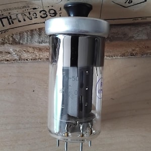 Può includere: Una valvola termoionica vintage con un involucro di vetro trasparente, una parte superiore nera e più pin alla base. Il tubo ha componenti interni e marcature, tra cui "9-50" e "OTK 76".