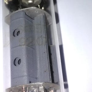 Puede incluir: Una vista cercana del interior de un tubo de vacío de vidrio. El tubo contiene un componente metálico gris con dos agujeros circulares. El componente está rodeado por una serie de finos cables metálicos.