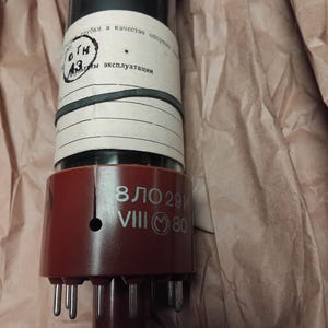 Puede incluir: Un tubo de vacío vintage con un cuerpo de vidrio oscuro y una base roja. La base tiene varios pines metálicos y el texto "ВЛО 29И VIII 80". El tubo está envuelto en papel y tiene una etiqueta con texto cirílico.