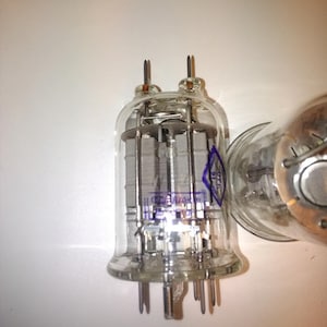 May include: A clear glass vacuum tube with multiple internal components and metal pins. The tube has a rounded base and two metal prongs at the top. A blue marking is visible on the glass. Another similar tube is partially visible in the background.