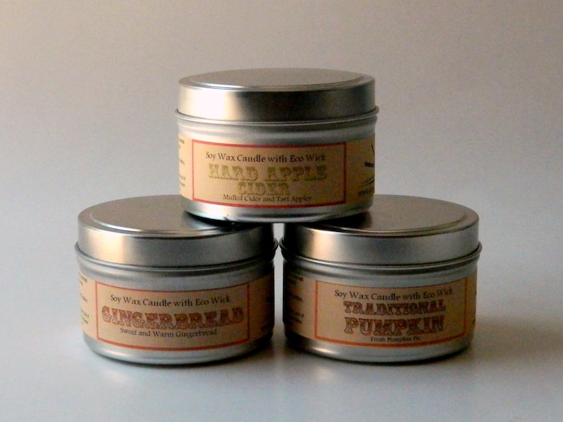May include: Three silver candle tins with labels. The labels are brown with gold text. The candles are labeled "Hard Apple Cider", "Gingerbread", and "Traditional Pumpkin".