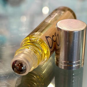 May include: A clear glass roller bottle with a silver cap and a label that says "DCO" in black lettering. The bottle is filled with a yellow liquid.