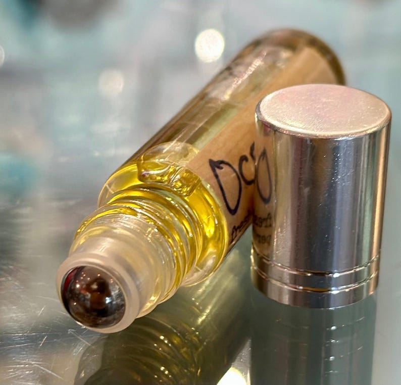 May include: A clear glass roller bottle with a silver cap and a label that says "DCO" in black lettering. The bottle is filled with a yellow liquid.