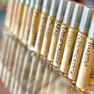 May include: A row of small glass bottles with silver roll-on tops. Each bottle has a label with a different name written on it, including "Tweedo", "Woodstock", "Palo", "Drift Away", "Man", "Ocean", "Riverwalk", and "Forestwalk".