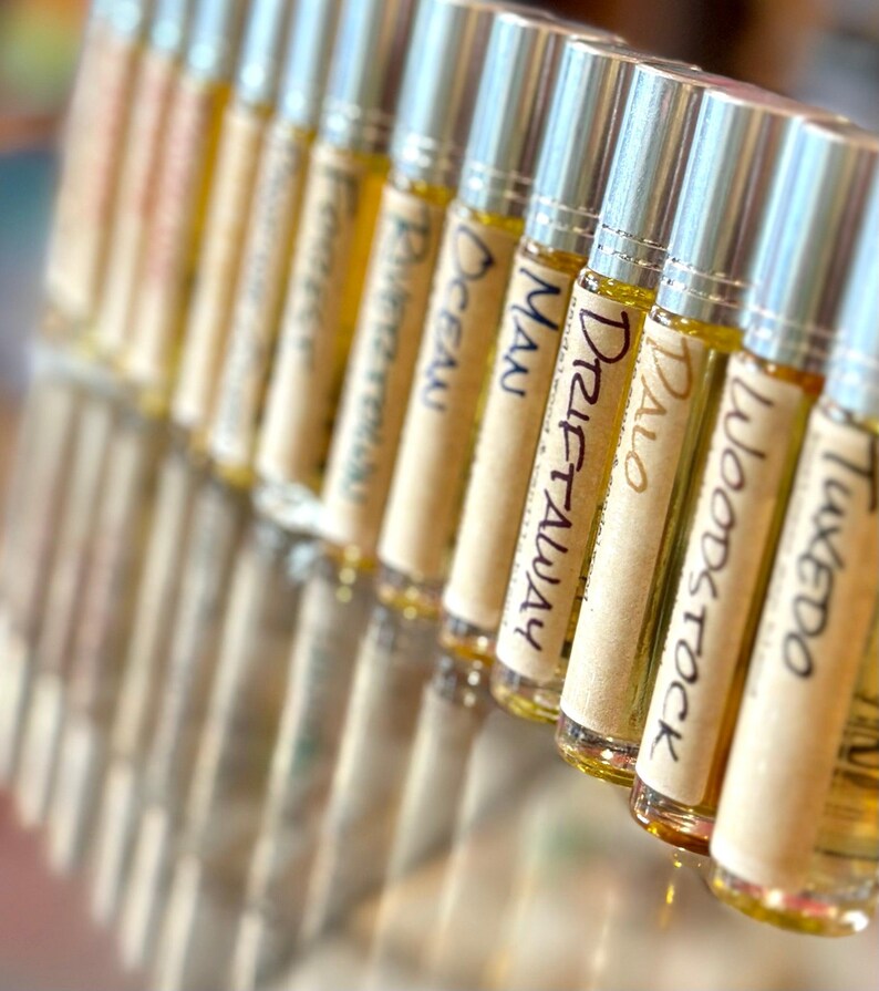 May include: A row of small glass bottles with silver roll-on tops. Each bottle has a label with a different name written on it, including "Tweedo", "Woodstock", "Palo", "Drift Away", "Man", "Ocean", "Riverwalk", and "Forestwalk".