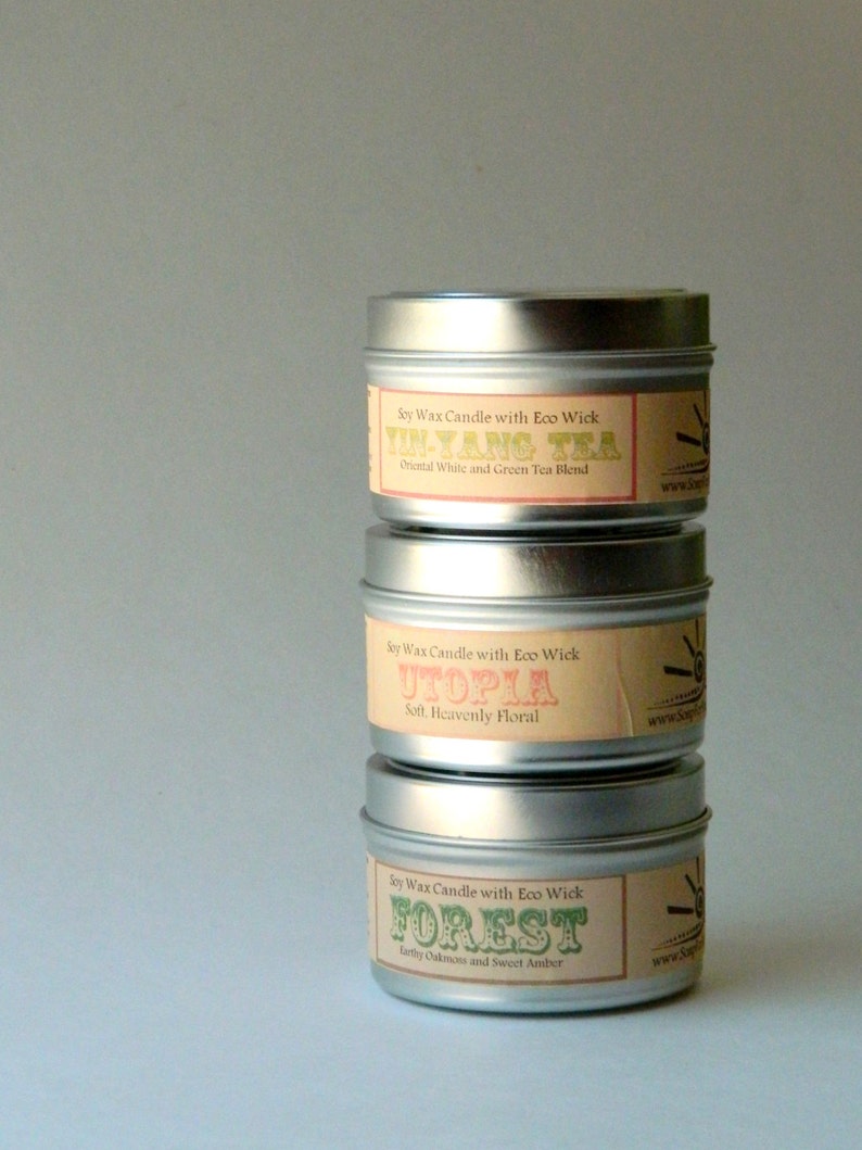 May include: Three silver candle tins with labels. The top tin has a label that says "Soy Wax Candle with Eco Wick Yin-Yang Tea Oriental White and Green Tea Blend". The middle tin has a label that says "Soy Wax Candle with Eco Wick Utopia Soft. Heavenly Floral". The bottom tin has a label that says "Soy Wax Candle with Eco Wick Forest Earthy Oakmoss and Sweet Amber".