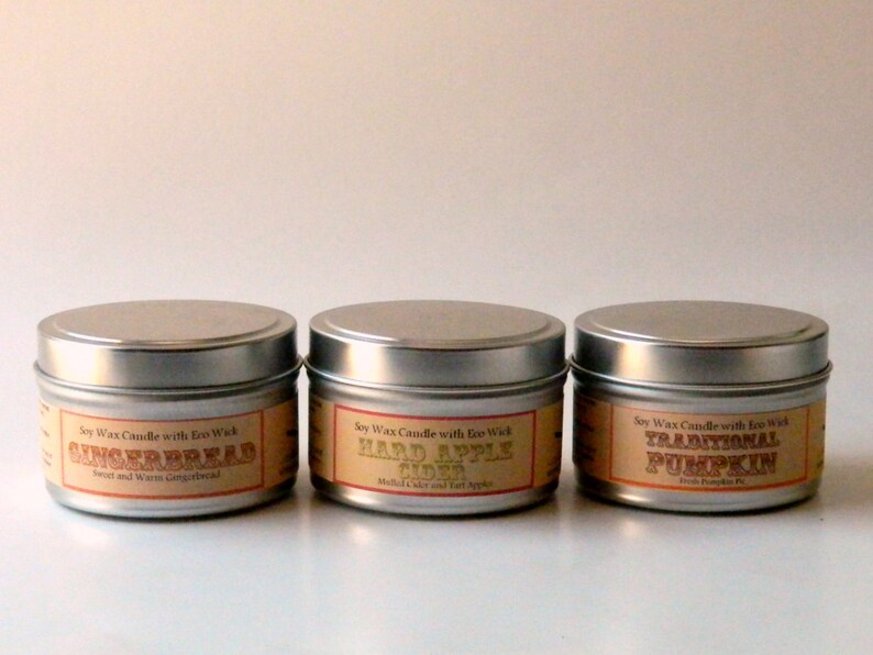 May include: Three silver candle tins with labels. The labels are orange and brown with white text. The labels read "Soy Wax Candle with Eco Wick" and the scents are "Gingerbread", "Hard Apple Cider", and "Traditional Pumpkin".