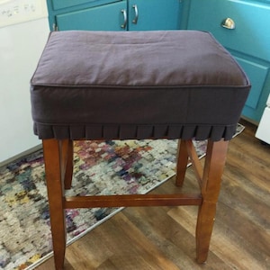 May include: A brown fabric chair cushion with a ruffled edge, covering a wooden bar stool with a brown finish.