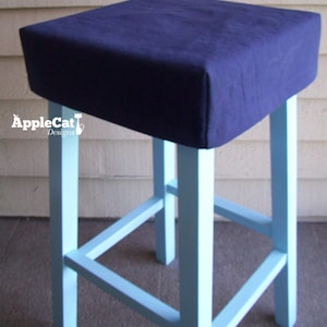 May include: A blue upholstered seat on a light blue wooden stool with a simple design. The stool has four legs and a single crossbar.