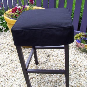 May include: A black square bar stool with a dark metal frame. The seat is a thick, padded cushion. The stool is outdoors on a bed of small white pebbles, with a purple fence and potted plants in the background.