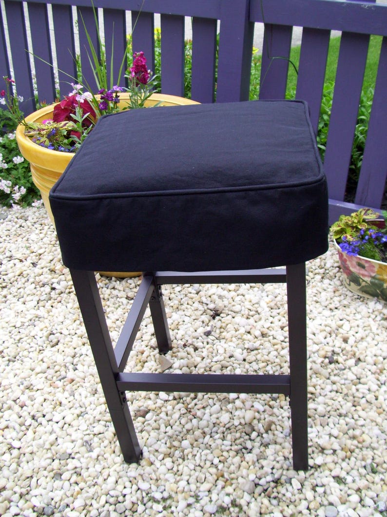 May include: A black square bar stool with a dark metal frame. The seat is a thick, padded cushion. The stool is outdoors on a bed of small white pebbles, with a purple fence and potted plants in the background.