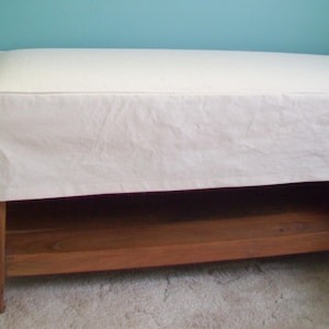 May include: A white upholstered bench with a wooden base and a shelf underneath. The bench is sitting on a light brown carpet.