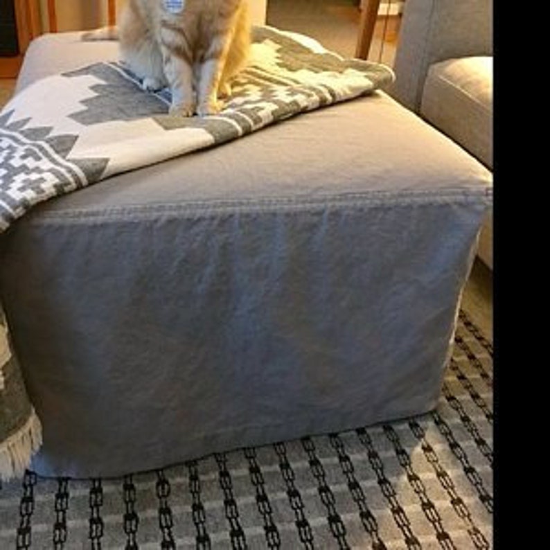 Pu&ograve; includere: Un pouf in lino grigio con una coperta a motivi bianchi e grigi sopra. Il pouf &egrave; su un tappeto a motivi bianchi e neri.