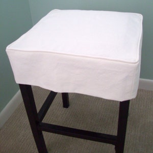 May include: A white square stool with a dark brown wooden frame. The stool has a white fabric seat cover with a piped edge. The legs are straight and the stool is on a light brown carpet.