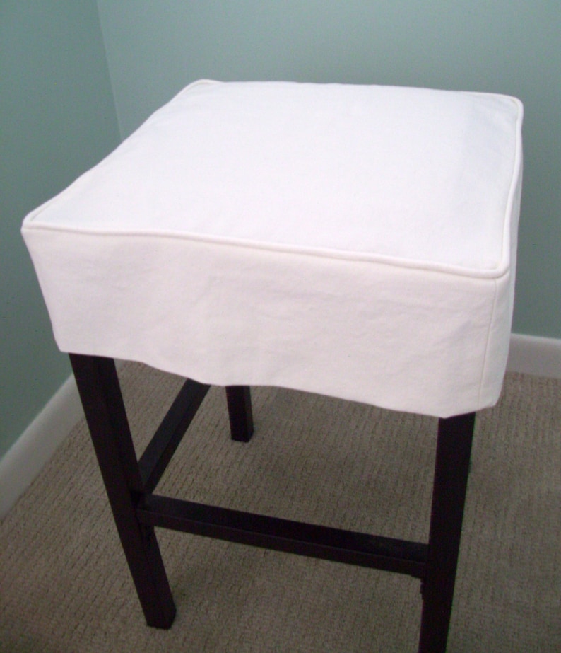 May include: A white square stool with a dark brown wooden frame. The stool has a white fabric seat cover with a piped edge. The legs are straight and the stool is on a light brown carpet.