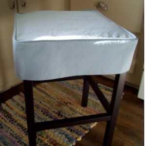 May include: A square, light blue upholstered bar stool with a dark brown wooden frame. The seat has a light blue cushion with a matching trim. The stool is set on a dark wooden floor, with a colorful rug visible below.