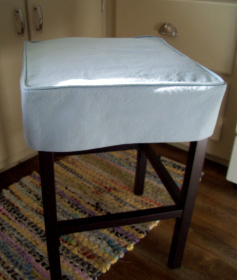 May include: A square, light blue upholstered bar stool with a dark brown wooden frame. The seat has a light blue cushion with a matching trim. The stool is set on a dark wooden floor, with a colorful rug visible below.
