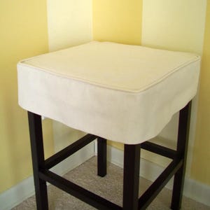 May include: A square bar stool with a cream-colored cushion and dark brown legs. The cushion has a piped edge. The stool's frame is made of dark wood, and the legs are connected by horizontal and diagonal supports.
