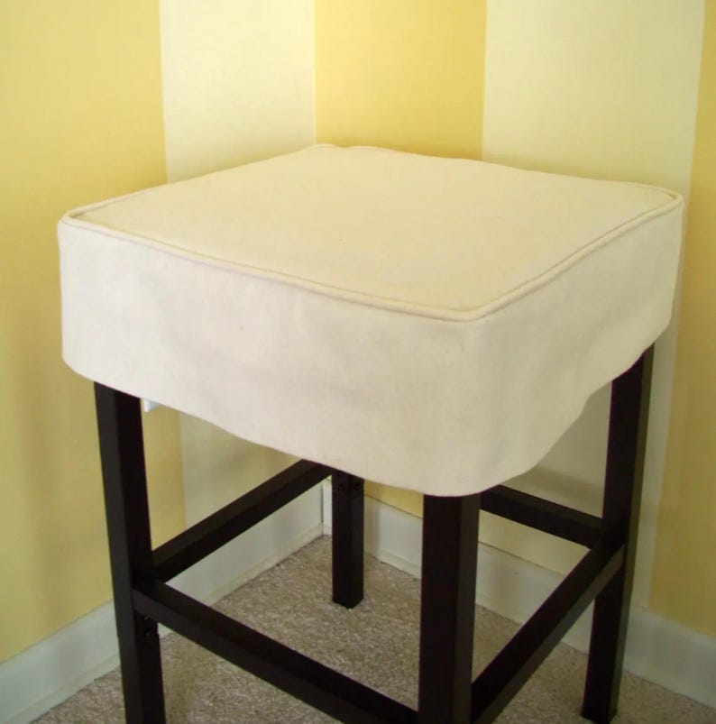 May include: A square bar stool with a cream-colored cushion and dark brown legs. The cushion has a piped edge. The stool's frame is made of dark wood, and the legs are connected by horizontal and diagonal supports.