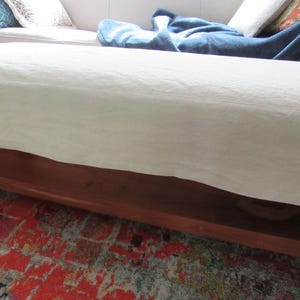 May include: A white linen ottoman with a wooden base and a shelf underneath. The ottoman is sitting on a colorful rug.