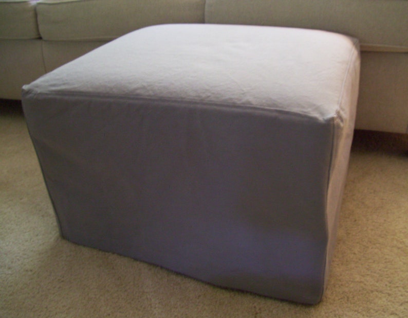 Pu&ograve; includere: Un pouf quadrato in tessuto grigio chiaro con un piano superiore liscio e piatto.