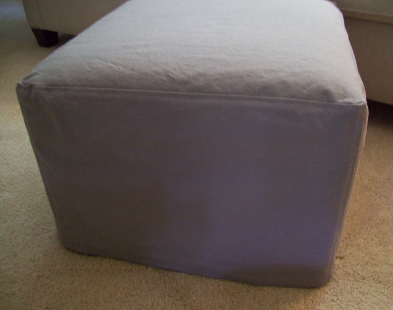 Pu&ograve; includere: Un pouf quadrato in tessuto grigio con un piano superiore liscio e piatto.