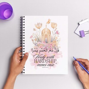Puede incluir: Cuaderno de espiral con un diseño floral y arquitectónico. La portada presenta un edificio dorado, flores y mariposas, con texto en árabe e inglés: "Verily with hardship comes ease."