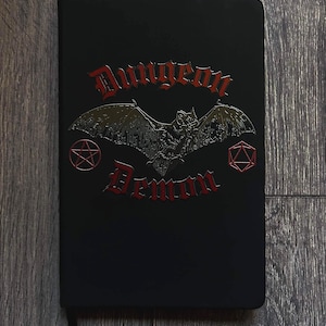 May include: Black notebook with the words "Dungeon Demon" in red gothic lettering. A bat with outstretched wings is centered above the text. A pentagram and a geometric shape are on either side of the bat.