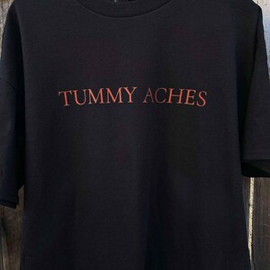 May include: Black t-shirt with the words "TUMMY ACHES" printed in orange across the front. The shirt is hanging on a wooden hanger. The text is in a bold, sans-serif font.
