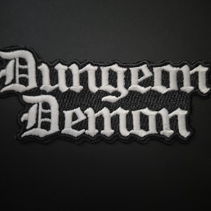 May include: Embroidered patch with the words "Dungeon Demon" in white gothic lettering on a black background. The patch has a black border and is likely for clothing or accessories.
