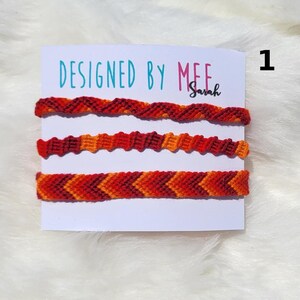 May include: Three handmade friendship bracelets in shades of red, orange, and burgundy are displayed on a white card. The card reads "DESIGNED BY MEE Sarah" in teal and pink. The bracelets feature various woven patterns.