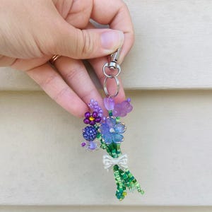 May include: A beaded keychain featuring a bouquet of flowers in shades of purple, blue, and green. The flowers are accented with a white bow and attached to a silver-colored clasp. The keychain is held against a light-colored background.