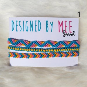 May include: Set of three friendship bracelets with a chevron pattern in pink, blue, and yellow. The bracelets are displayed on a white card with the text "DESIGNED BY MEE Sarah".