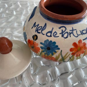 Può includere: Un piccolo vaso rotondo in ceramica con coperchio, dipinto a mano con fiori colorati e le parole "Mel de Portugal". Il vaso ha un bordo marrone e un pomello marrone abbinato sul coperchio. Lo sfondo è una superficie trasparente e strutturata.