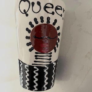 Puede incluir: Un vaso de cerámica blanco con la palabra "Queen" en escritura negra. El vaso presenta un diseño rojo en forma de sol con detalles en negro y un patrón ondulado en blanco y negro en la base. El vaso mide unos 15 cm de alto.