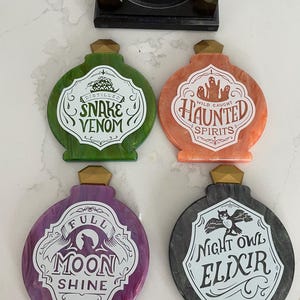 Puede incluir: Cuatro posavasos con forma de botella con acabados acrílicos jaspeados en verde, naranja, morado y negro. Cada uno tiene un tapón dorado y una etiqueta blanca con texto en negro: Distilled Snake Venom, Wild Caught Haunted Spirits, Full Moon Shine y Night Owl Elixir.