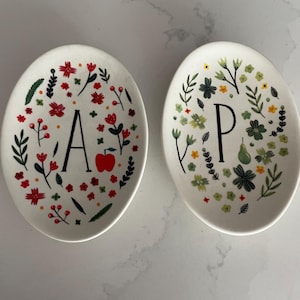 Puede incluir: Dos platos de cer&aacute;mica ovalados blancos. Uno presenta la letra "A" con flores rojas, hojas verdes y una manzana roja. El otro tiene la letra "P" con dise&ntilde;os florales verdes y amarillos. Art&iacute;culos de cocina decorativos.