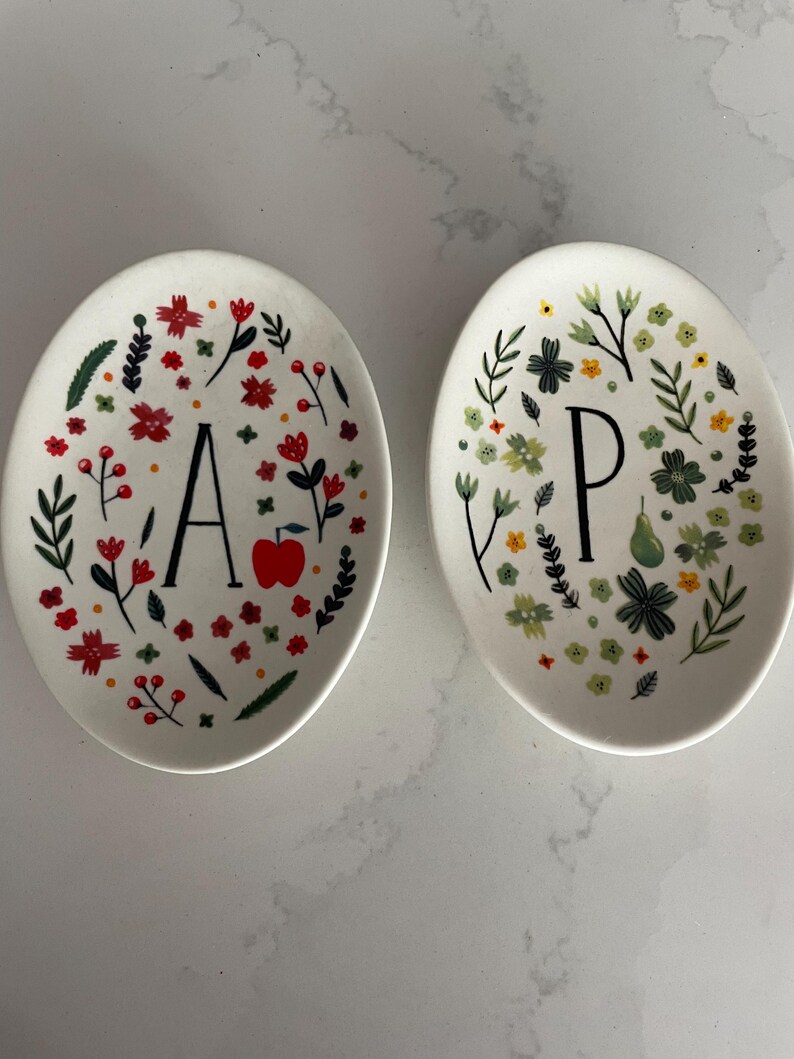 Puede incluir: Dos platos de cer&aacute;mica ovalados blancos. Uno presenta la letra "A" con flores rojas, hojas verdes y una manzana roja. El otro tiene la letra "P" con dise&ntilde;os florales verdes y amarillos. Art&iacute;culos de cocina decorativos.