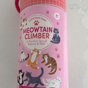 May include: A pink cylinder-shaped toy package with a red handle features the text "MEOWTAIN CLIMBER" and illustrations of stacking cats. The package includes the text "A Purrfect Test of Balance & Skill" and "INCLUDES 12 STACKING CATS".