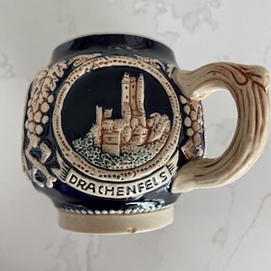 Puede incluir: Una pequeña jarra de cerveza de cerámica con un cuerpo azul oscuro y un asa de color beige claro. La jarra presenta un relieve detallado de una escena de castillo dentro de un marco circular, con la palabra "DRACHENFELS" debajo. Uvas y vides decoran los lados.