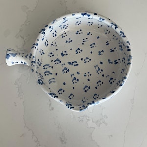 Könnte beinhalten: Weißes Keramik-Serviergeschirr mit Griff, verziert mit blauen Blumenmustern. Das Geschirr hat eine runde Form mit einem gewellten Rand. Der Griff ist ebenfalls weiß mit blauen Akzenten. Auf einer weißen Marmoroberfläche.
