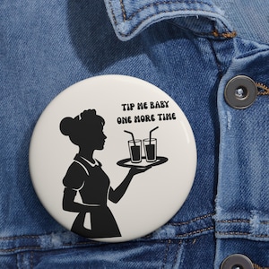 May include: A white button featuring a black silhouette of a waitress holding a tray with two drinks. The text "TIP ME BABY ONE MORE TIME" is printed above the drinks. The button is set against a blue denim background.