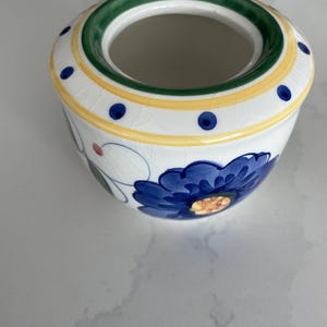 Peut inclure: Un sucrier en céramique blanche orné d'un motif de fleur bleue peint à la main avec des accents jaunes et verts. Le bord est décoré d'une bande verte et d'une rayure jaune avec des points bleus. L'émail présente une finition craquelée visible.